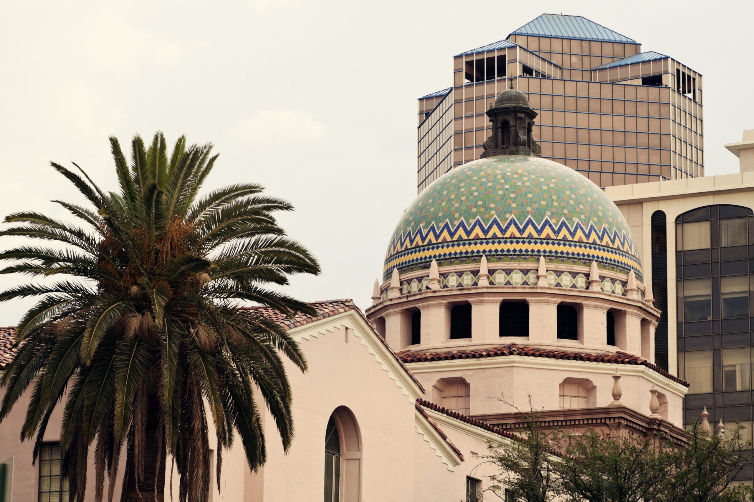 A domed historic building with colorful tiles stands in front of modern office buildings and a palm tree.